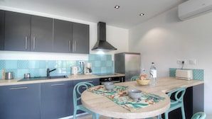 Une cuisine de taille réduite, avec des meubles de couleur sombre, un réfrigérateur en acier inoxydable, une machine à café et une table agrémentée d’un set de table à motifs floraux.