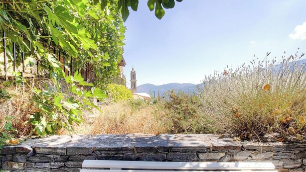 A stone bench in a garden with a stone wall, greenery, and a distant view of a bell tower. - A Torra - AITI - Gîte (AITI)