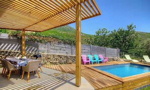 Una terraza de madera con piscina, mobiliario exterior y un muro de piedra.