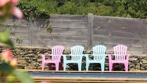 Tres sillas Adirondack en tonos rosa, blanco y azul, dispuestas en fila frente a un muro de piedra con una valla de madera al fondo.
