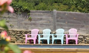 Three Adirondack chairs in pink, white, and blue colors, arranged in a row against a stone wall with a wooden fence in the background. - U Sognu - CAMPITELLO (CAMPITELLO)