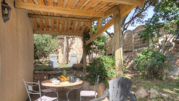 Een overdekt terras met een tafel en stoelen, omringd door natuurlijke rotsformaties en groen.