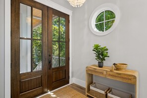 Una puerta doble de madera con paneles de vidrio, un ventanal redondo, una mesa consola con cestas y una planta en maceta.