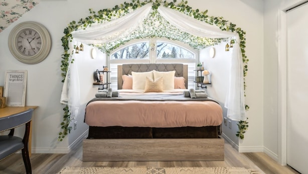 A bedroom with a canopy bed, a clock, a bedside table, and a view of trees outside. - Central 3BR Home | Sleeps 8, 4 Beds + 2 Bathrooms (Calgary)