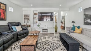 A modern living room with a black leather sofa, a wooden coffee table, and a grey sofa with colorful cushions. A large TV is mounted on the wall, and there are shelves with decorative items and a vase. - Central 3BR Home | Sleeps 8, 4 Beds + 2 Bathrooms (Calgary)