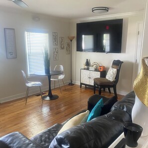 Ein Wohnzimmer mit schwarzem Ledersofa, einem Esstisch mit Stühlen, einem an der Wand montierten Fernseher und einer Stehlampe.