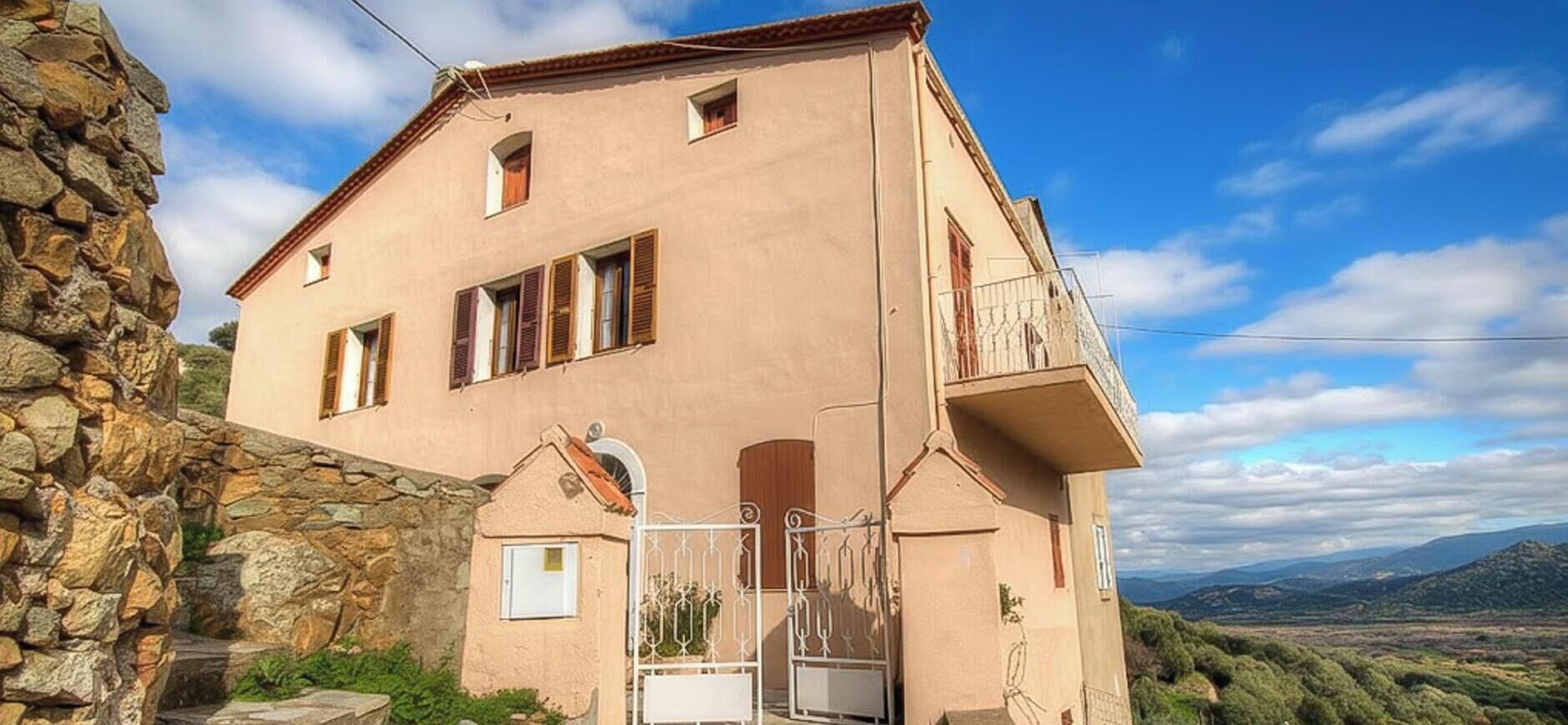 Un bâtiment à deux étages, avec un mur en pierre d’un côté, un balcon à l’étage supérieur et une vue sur la campagne.