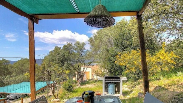Ein überdachter Außenbereich mit einer für zwei Personen gedeckten Tafel, einer hängenden Leuchte und Blick auf eine natürliche Landschaft mit Bäumen und einem Gebäude im Hintergrund.