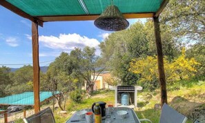 A covered outdoor seating area with a table set for two, a hanging light fixture, and a view of a natural landscape with trees and a building in the background. - A Machja - FIGARI - Gîte (FIGARI)