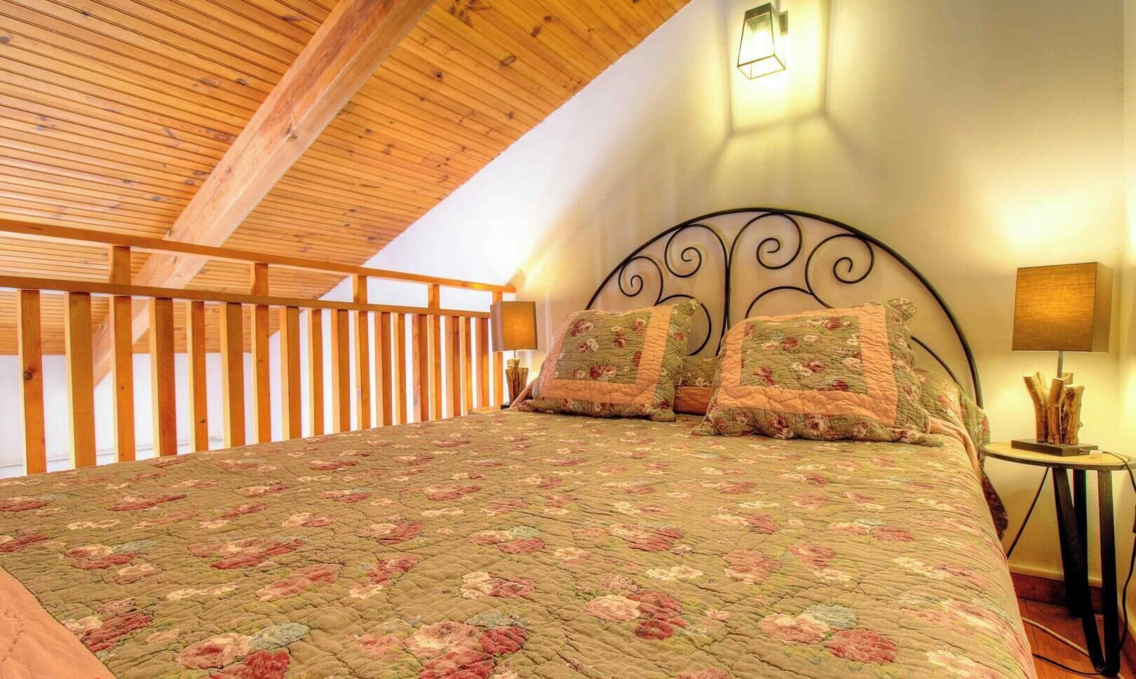 A bedroom with a wooden ceiling, a bed with floral bedding, a metal headboard, and a nightstand with a lamp.