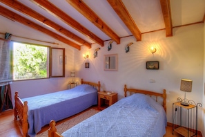 Ein Zimmer mit zwei Holzbetten, einem Fenster mit Blick auf Grünflächen, einem Spiegel, Wandleuchten und einem kleinen Tisch mit Lampe.