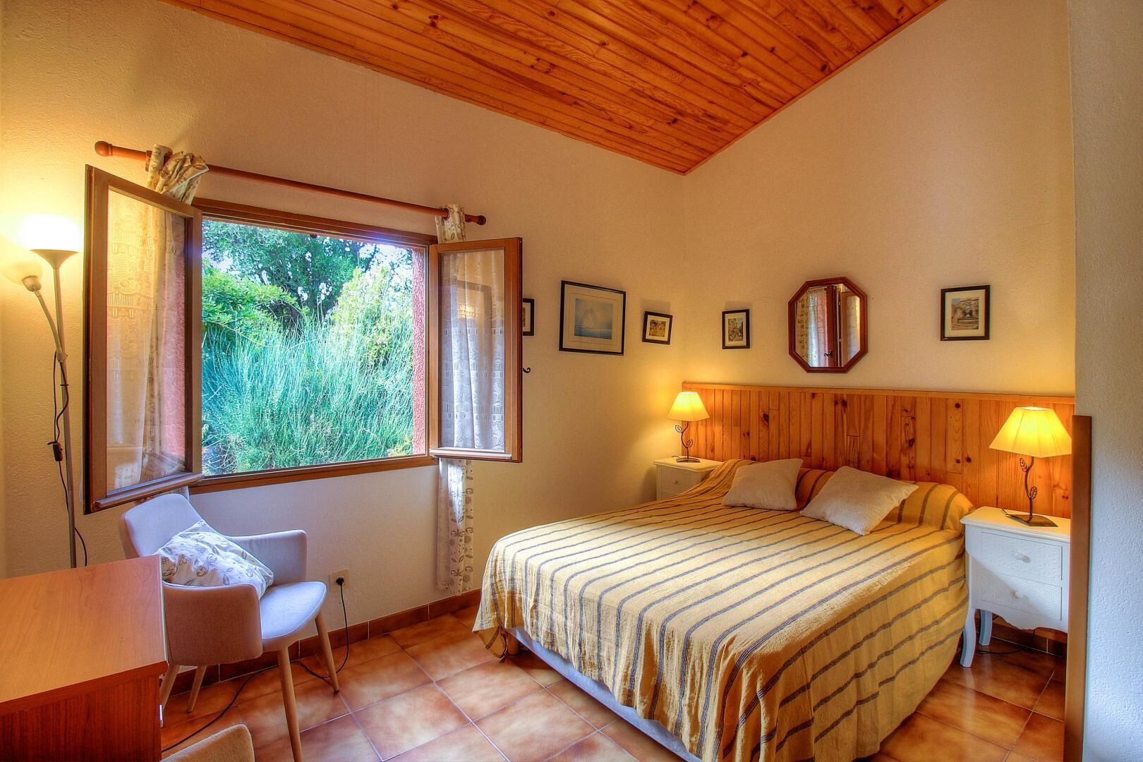A bedroom with a bed, two bedside tables, a chair, a desk, a window with curtains, and a wooden ceiling.