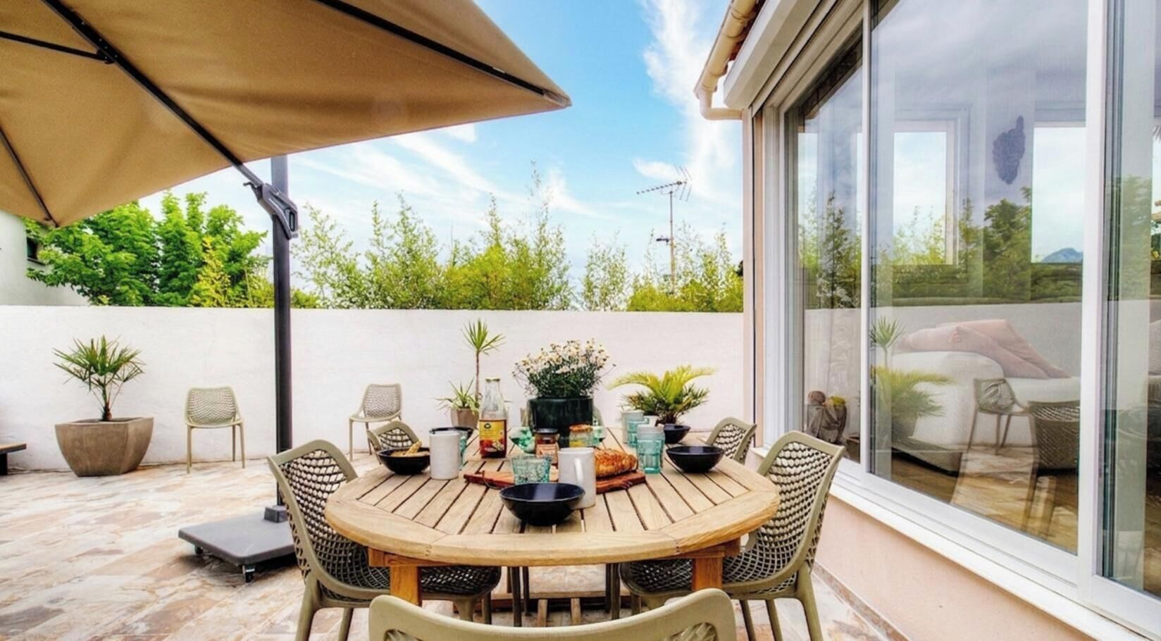 A round wooden table with chairs, a beige umbrella, and potted plants.