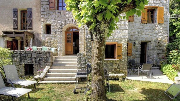 Un edificio de piedra con puerta de madera, mesas al aire libre y un árbol en primer plano.