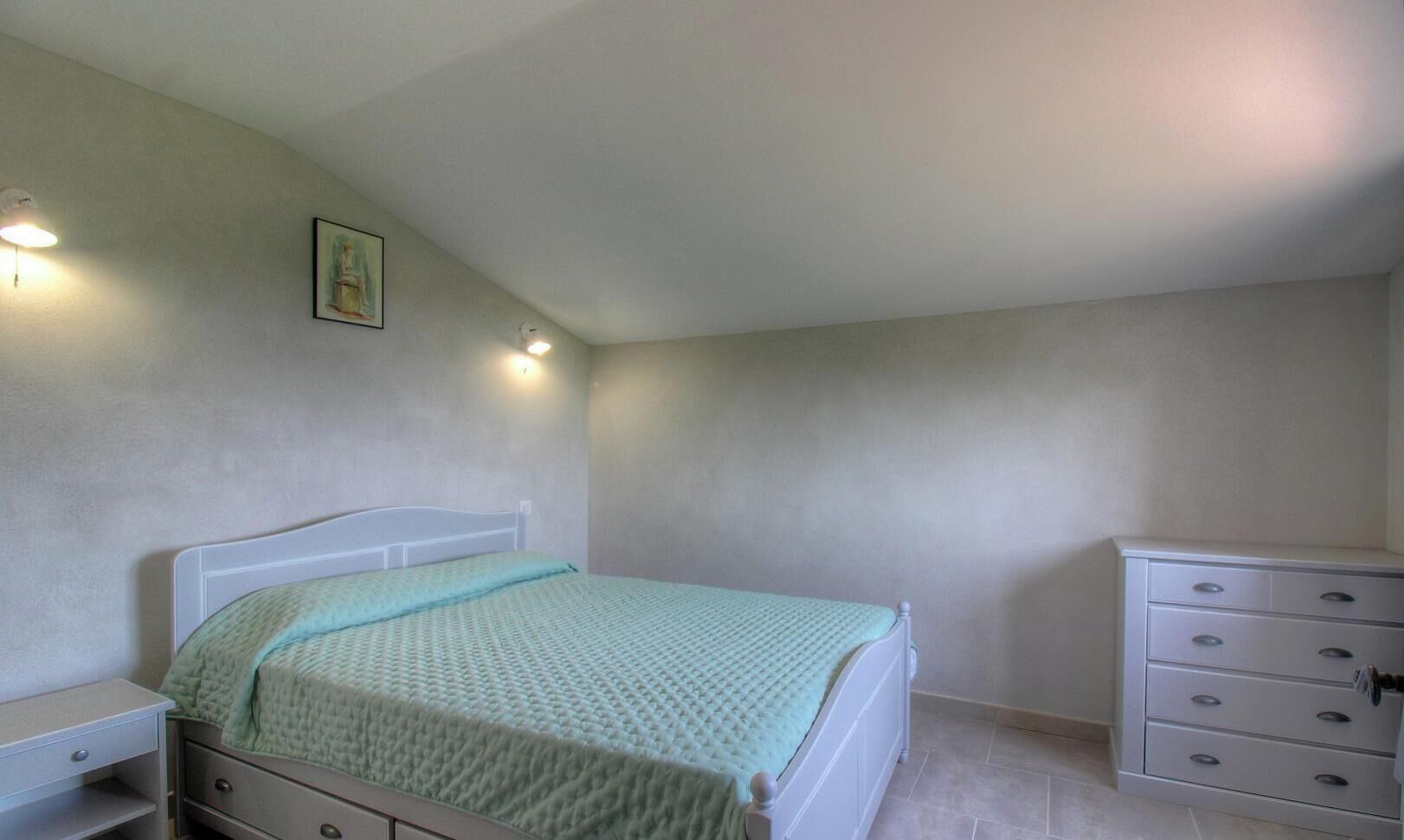 A bedroom with a bed, a dresser, a nightstand, and a wall-mounted light fixture.