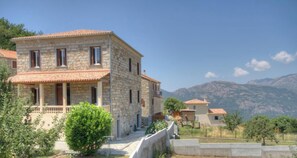 A stone building with a balcony, surrounded by greenery and mountains in the background. - A Foce - ALTAGENE - Gîte (ALTAGENE)