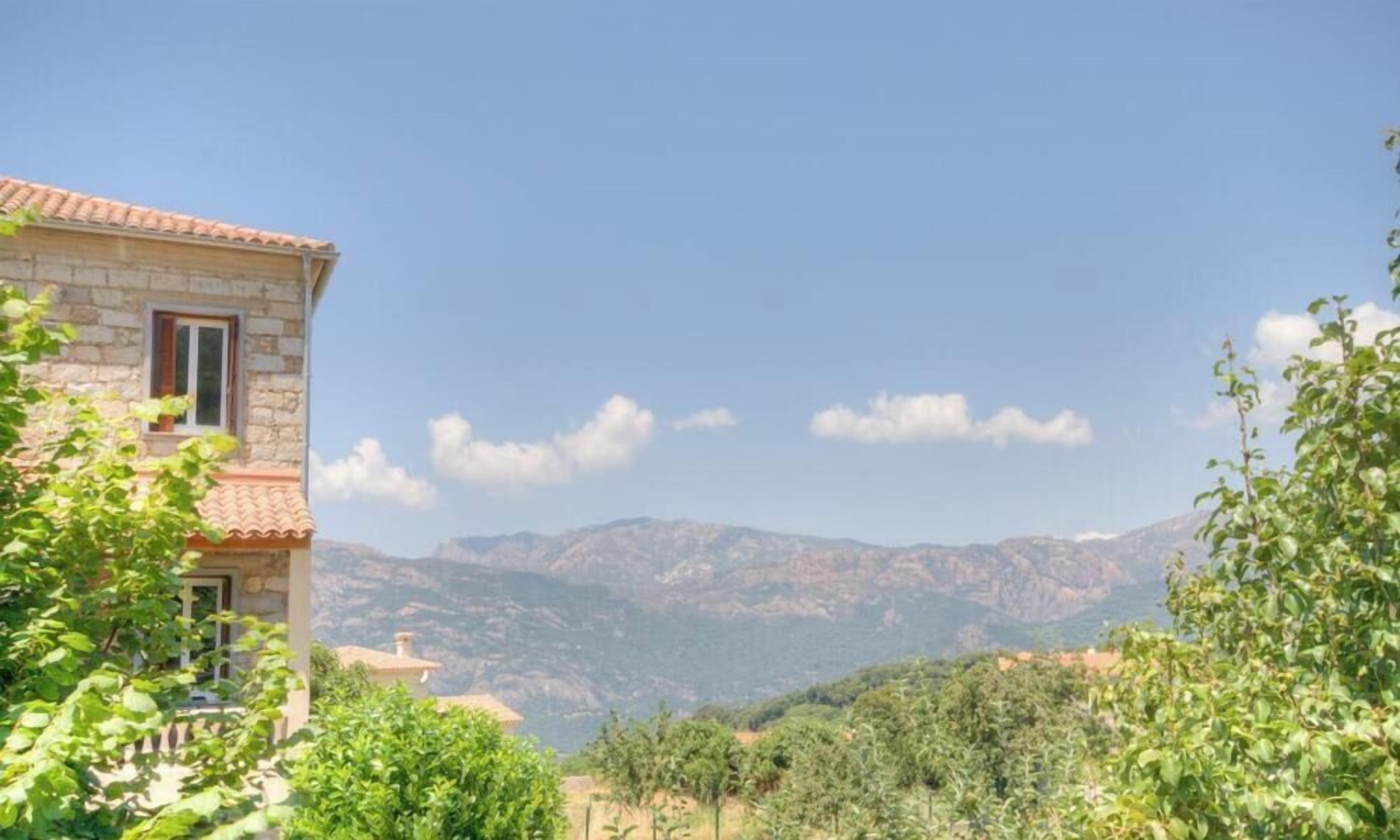 A stone building with a red-tiled roof, surrounded by greenery and mountains in the background.
