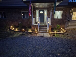 Una casa con fachada de ladrillo, un porche con escalones, una bandera y un jardín con luces.