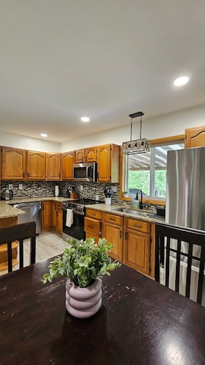 A kitchen with wooden cabinets, stainless steel appliances, a granite countertop, and a dining table with a vase of flowers. - Poconos Group Getaway: Hot Tub, Firepit& Pet Friendly (Lakeville)