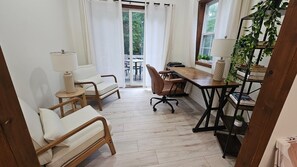 Una habitación con piso de madera, un escritorio con una silla, un sofá, una lámpara y una ventana con cortinas.