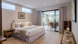 Ein Schlafzimmer mit einem großen Bett, einem Nachttisch, einer Essecke mit Tisch und Stühlen und Blick ins Freie durch eine Glastür.