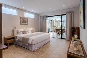 Ein Schlafzimmer mit einem großen Bett, einem Nachttisch, einer Essecke mit Tisch und Stühlen und Blick ins Freie durch eine Glastür.