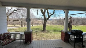 A covered porch with a white ceiling, a white fence, a wooden deck, a brick column, a metal bench, and a grill. - Peaceful country farmhouse, 2 bedrooms-all amenities for your comfort ! (Dansville)