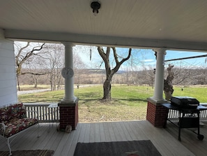 A covered porch with a white ceiling, a white fence, a wooden deck, a brick column, a metal bench, and a grill.