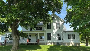 A two-story house with a porch, a large tree in front, and a clear sky. - Peaceful country farmhouse, 2 bedrooms-all amenities for your comfort ! (Dansville)