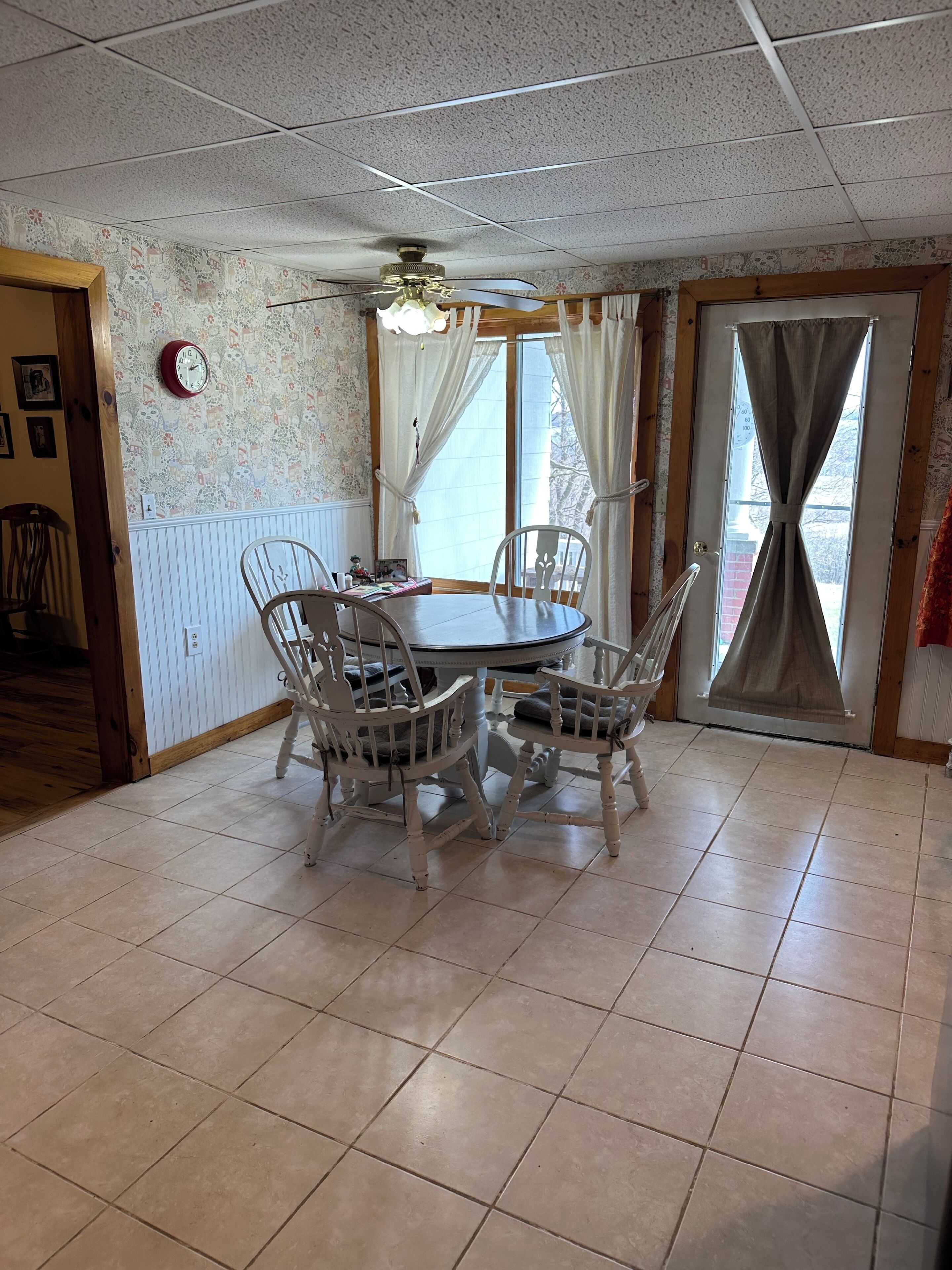 Un coin repas avec une table ronde et quatre chaises, un ventilateur de plafond, une horloge et une fenêtre avec des rideaux.