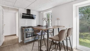 A kitchen with a gas stove, a stainless steel oven, a black range hood, a wooden table, two bar stools, and a view of greenery through a large window. - 4 Star Holiday Home in Spottrup (Spøttrup)