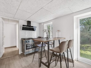 A kitchen with a gas stove, a stainless steel oven, a black range hood, a wooden table, two bar stools, and a view of greenery through a large window. - 4 Star Holiday Home in Spottrup (Spøttrup)