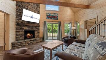 A log cabin interior with a stone fireplace, wooden ceiling beams, a sofa, and a dining table.