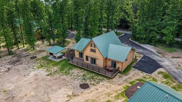 A two-story log cabin with a green metal roof, surrounded by a wooded area.