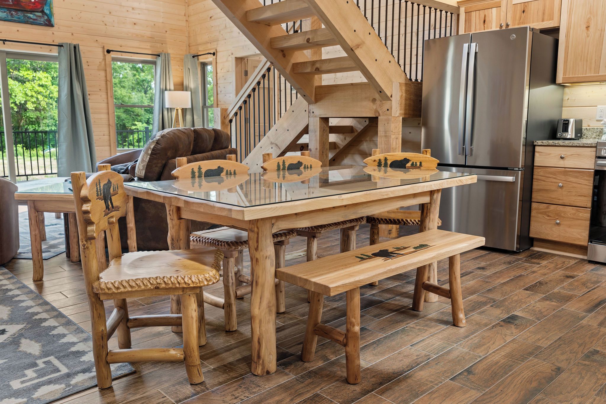 A wooden interior with a glass-top dining table, chairs, a bench, a kitchen area with a refrigerator, and a staircase.