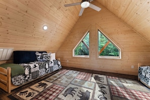 A cozy attic bedroom with a sloped ceiling, a bed with patterned bedding, a window with a view of trees, and a ceiling fan. - Authentic Log Cabin w Private Hot Tub_Near Big Cedar & Thunder Ridge_Free Tickets_Bonus Loft (Ridgedale)