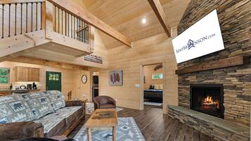 A cozy cabin interior with a wooden staircase, a fireplace, a sofa, a dining table, and a kitchen area.