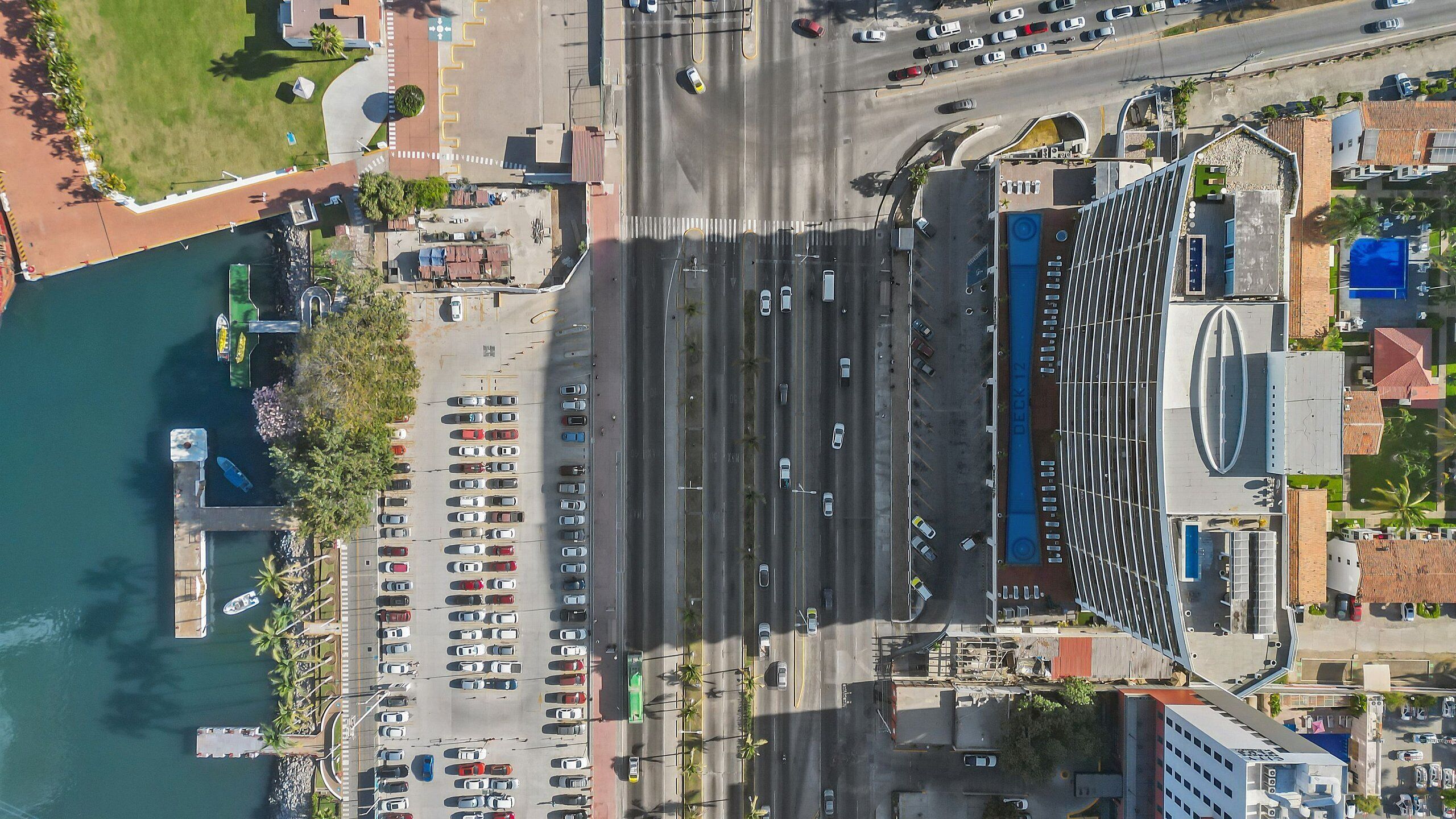 Luchtbeeld van een kuststedelijk gebied met een jachthaven, parkeerplaatsen en een groot gebouw met een opvallend dak.