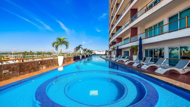 A swimming pool with lounge chairs and palm trees, surrounded by a building with balconies. - Deck12 2BR Premium Pool Access Parking (Puerto Vallarta)