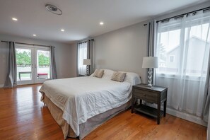 Ein Schlafzimmer mit einem großen Bett, einem Nachttisch, einer Lampe und Blick durch ein Fenster.