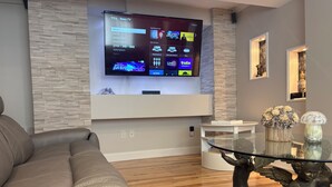 Uma sala de estar moderna com uma TV montada exibindo a interface de um serviço de streaming, um sofá de couro, uma mesa de centro de vidro com objetos decorativos e um tapete estampado.