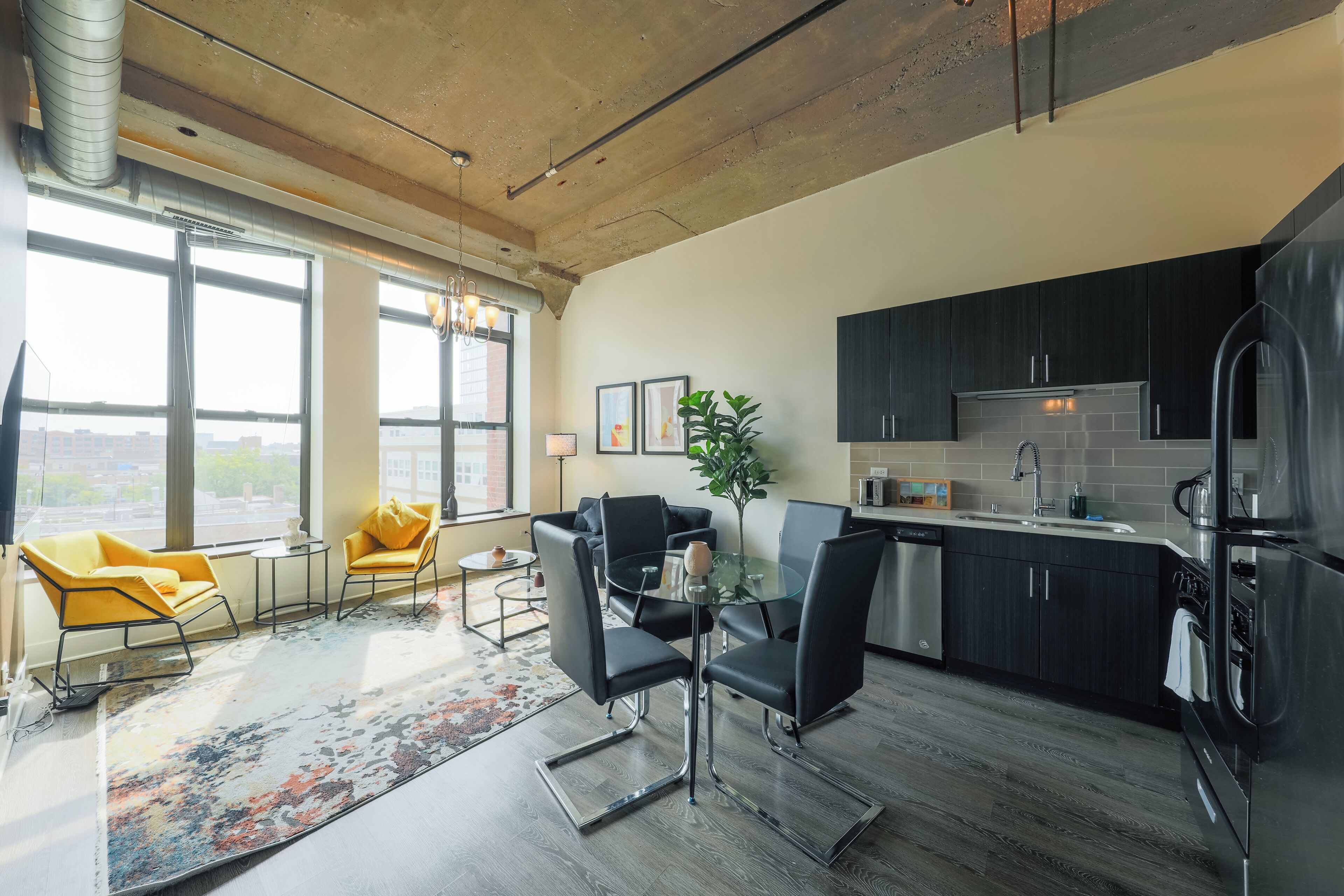 A modern kitchen with dark cabinets, a stainless steel refrigerator, and a dining area with a glass table and black chairs.