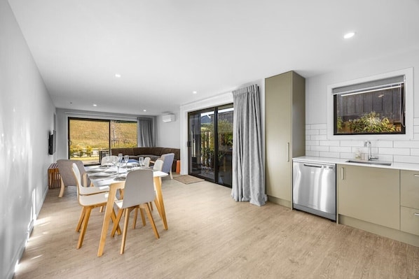 A modern kitchen with a dishwasher, a dining area with a table and chairs, and a sliding glass door leading to a balcony. - 3-bed in Northlake | Bikes, Boards & Wānaka Bliss (Wānaka)