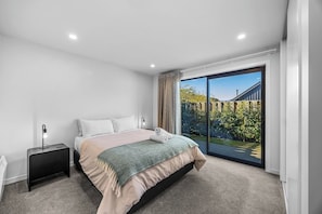 A modern bedroom with a large bed, a bedside table, a lamp, and a view of a garden through a sliding glass door. - 3-bed in Northlake | Bikes, Boards & Wānaka Bliss (Wānaka)