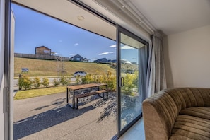 A balcony with a wooden table and chairs, a sofa, and a view of a hilly landscape with a building and a car. - 3-bed in Northlake | Bikes, Boards & Wānaka Bliss (Wānaka)