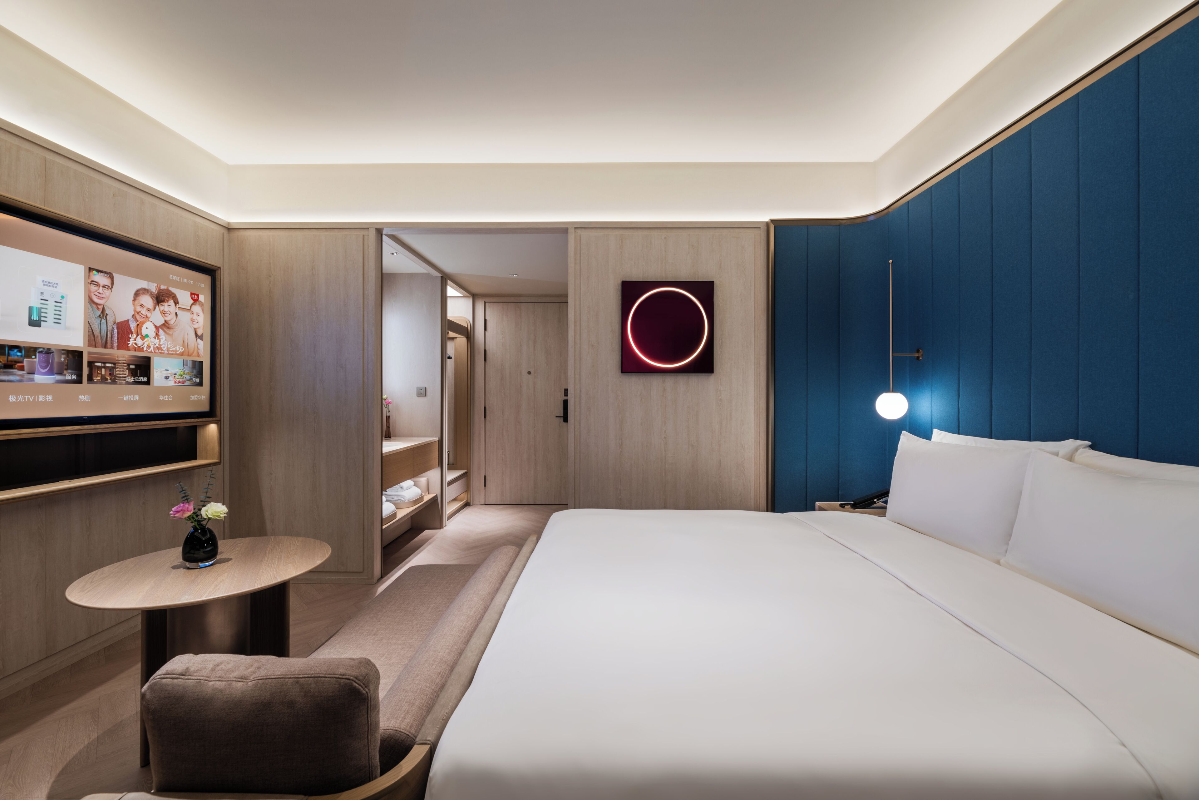 A modern hotel room with a large bed, a desk with a computer, a small seating area, and a wall-mounted TV.