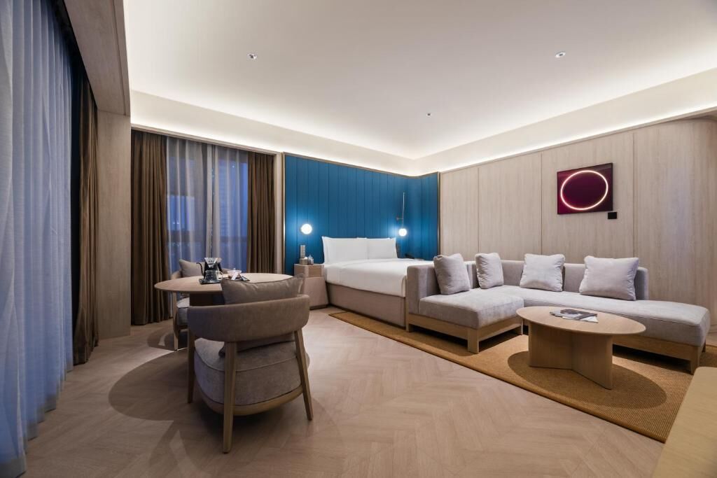 A modern hotel room with a bed, sofa, dining table, and a small coffee table.
