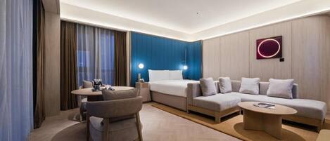 A modern hotel room with a bed, sofa, dining table, and a small coffee table.