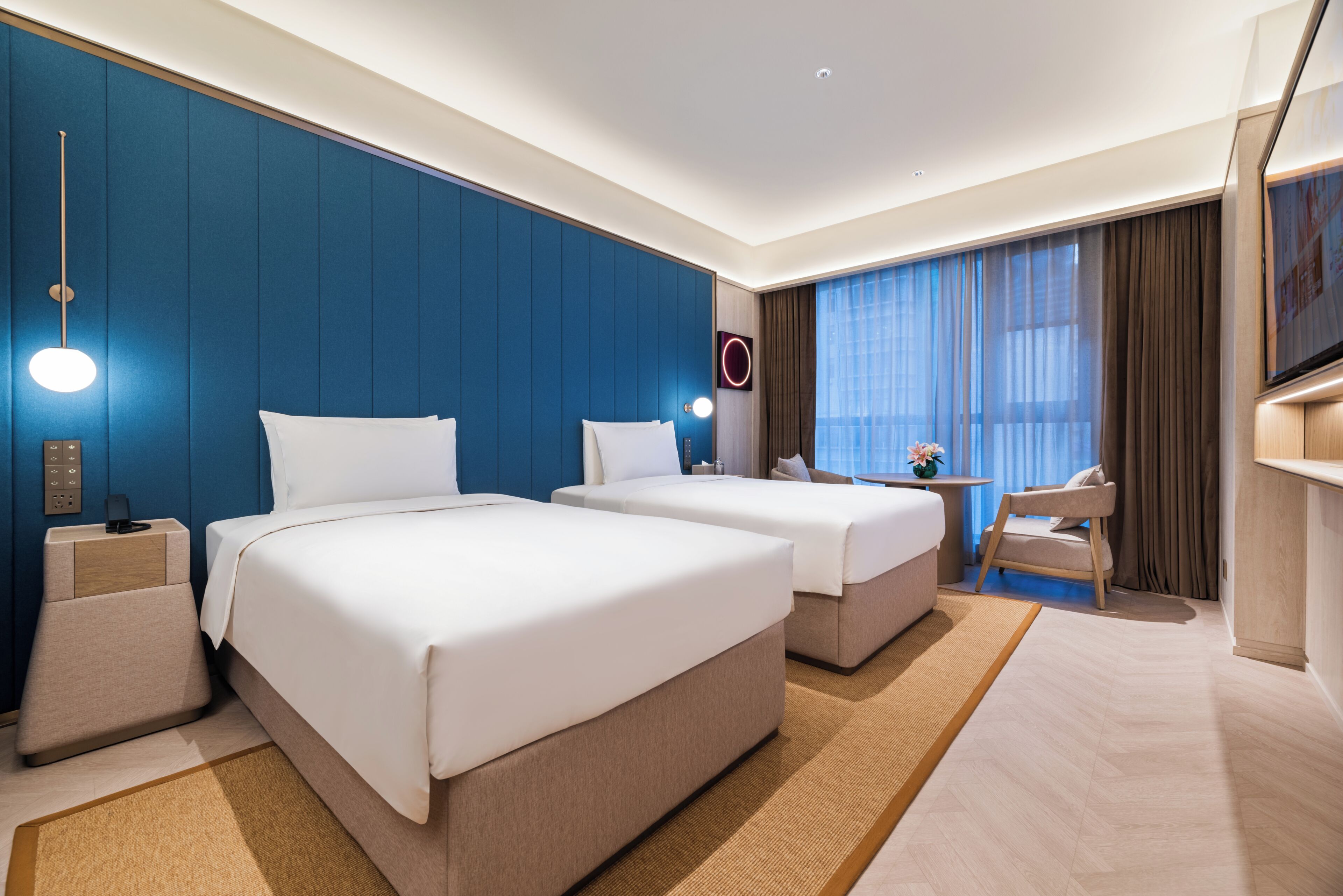 A hotel room with two beds, a desk, a chair, and a large window with curtains.