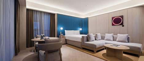 A modern hotel room with a bed, sofa, round coffee table, and a small dining table with chairs.